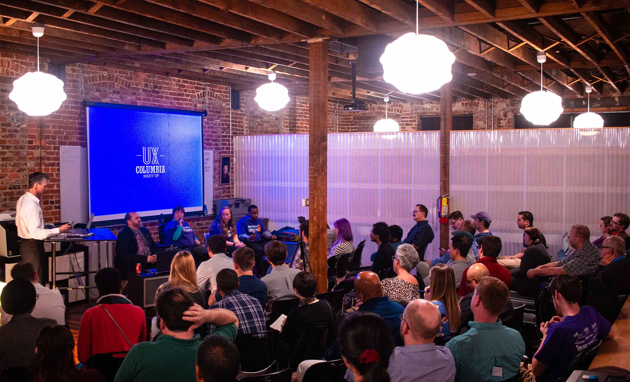 An audience of around thirty people sit looking towards a screen. In front of the screen stands a moderator and four panelists.